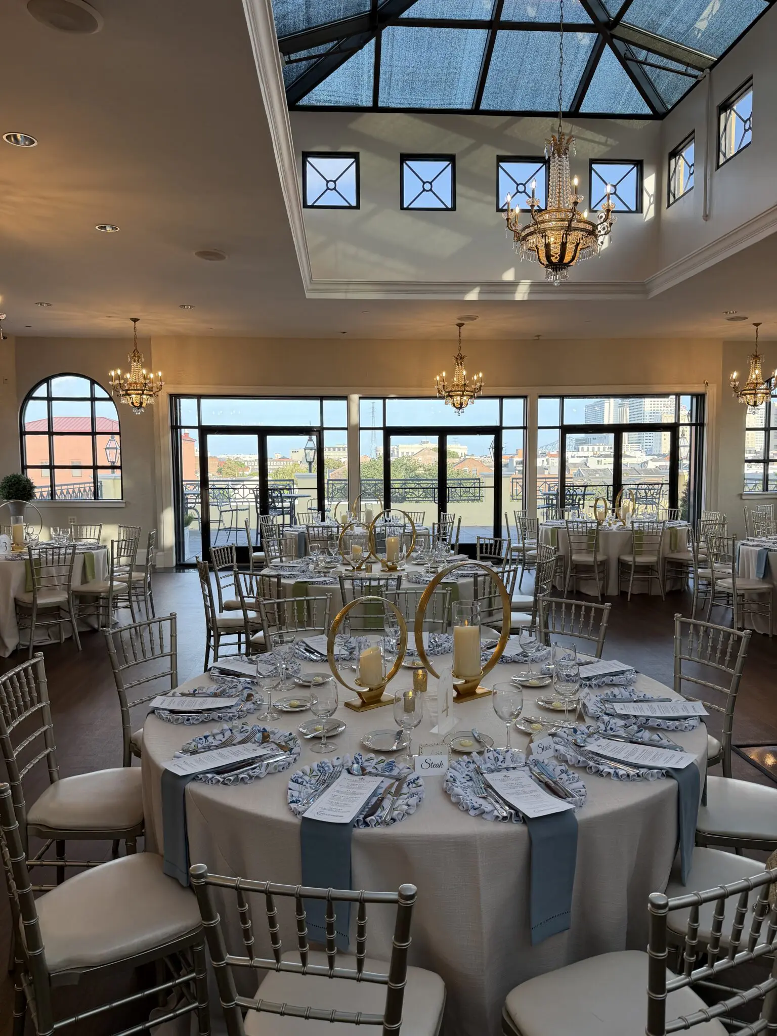 Wedding reception setup at The MISI by Messina’s in New Orleans, featuring round dining tables, blue-and-white place settings, candles, and a glass-enclosed rooftop view.